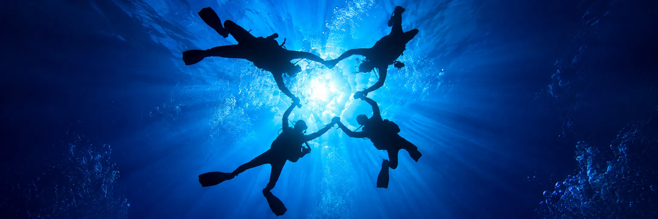 Four scuba diver silhouettes beneath the sunlit blue ocean surface